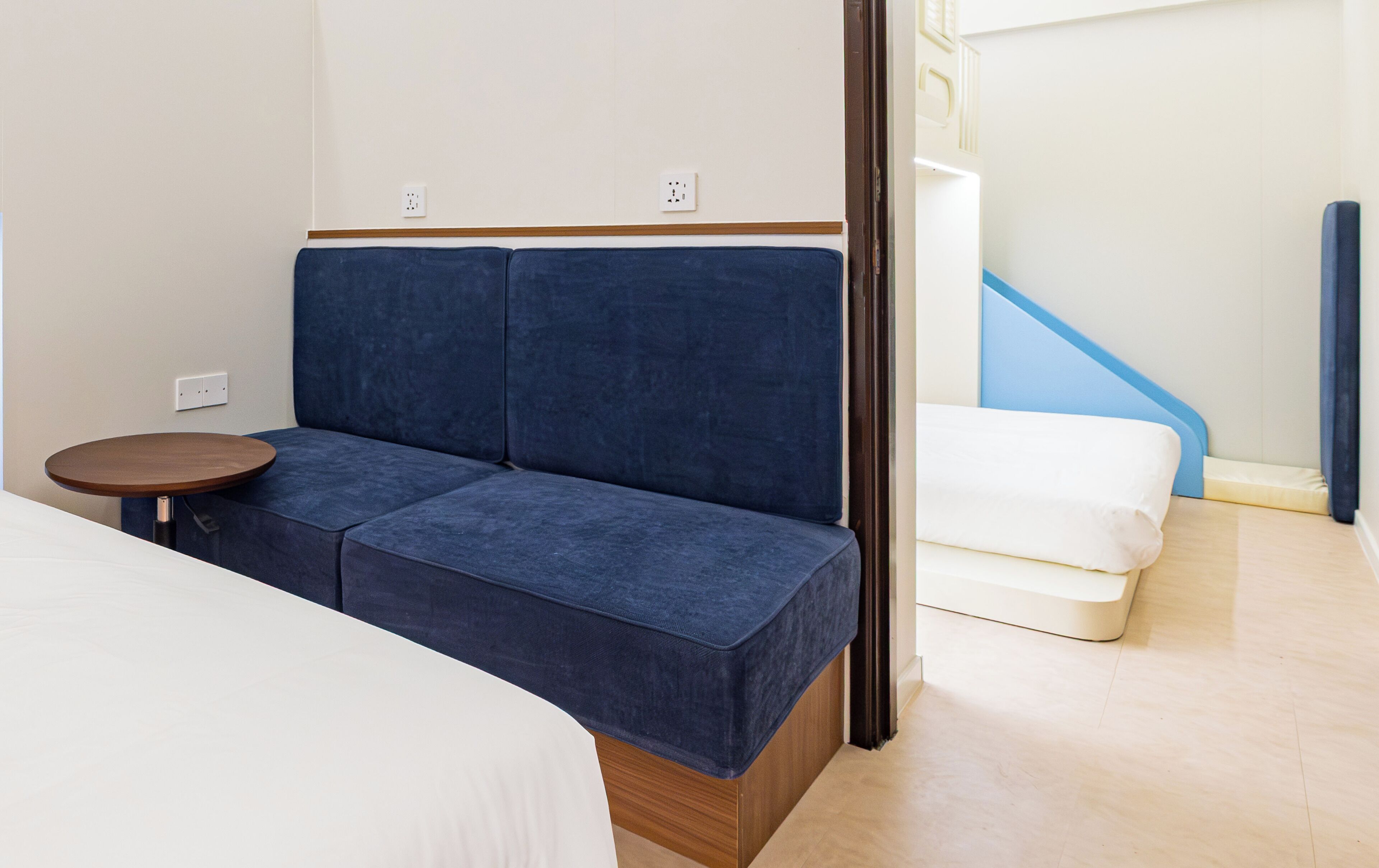 Habitación de hotel con sofá azul, una mesita redonda pequeña, cama blanca y una escalera con detalles azules.