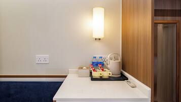 Habitación de hotel con un mostrador blanco, una lámpara de pared, una tetera, una cafetera y una caja de pañuelos.
