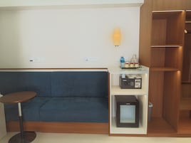 A room with a blue sofa, a small round table, a microwave, and a shelf with a water bottle and cups.