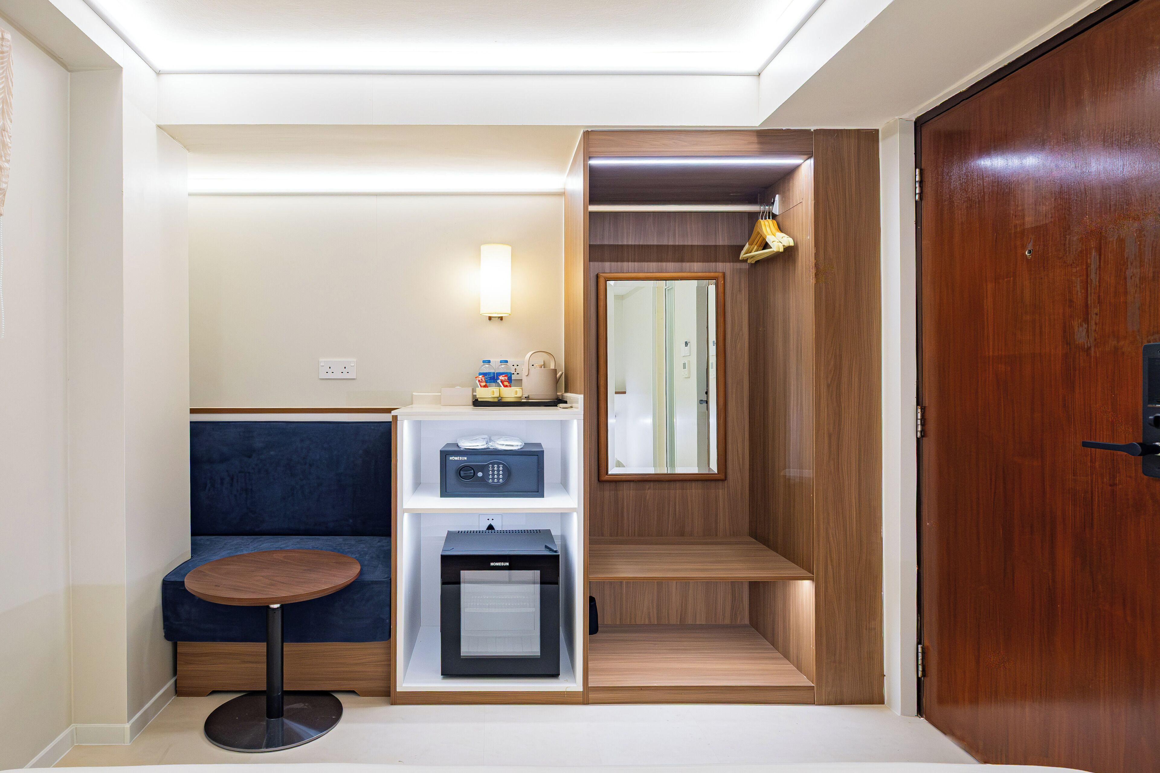 Ein kleines Hotelzimmer mit Sitzgelegenheit, einer Minibar, einem Spiegel und einer Holztür.