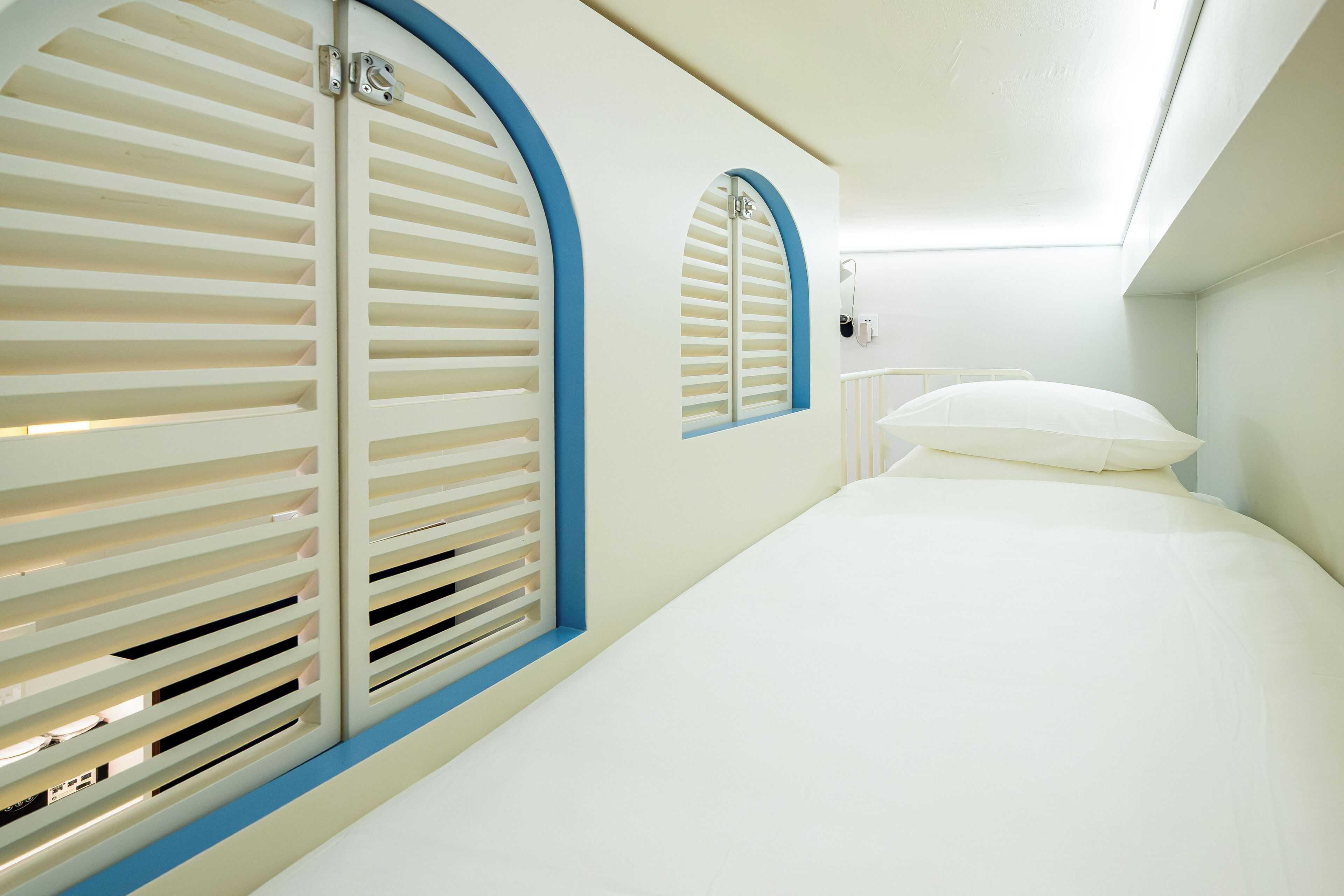 Una habitación con cama, ventana con persianas y una lámpara fijada a la pared.