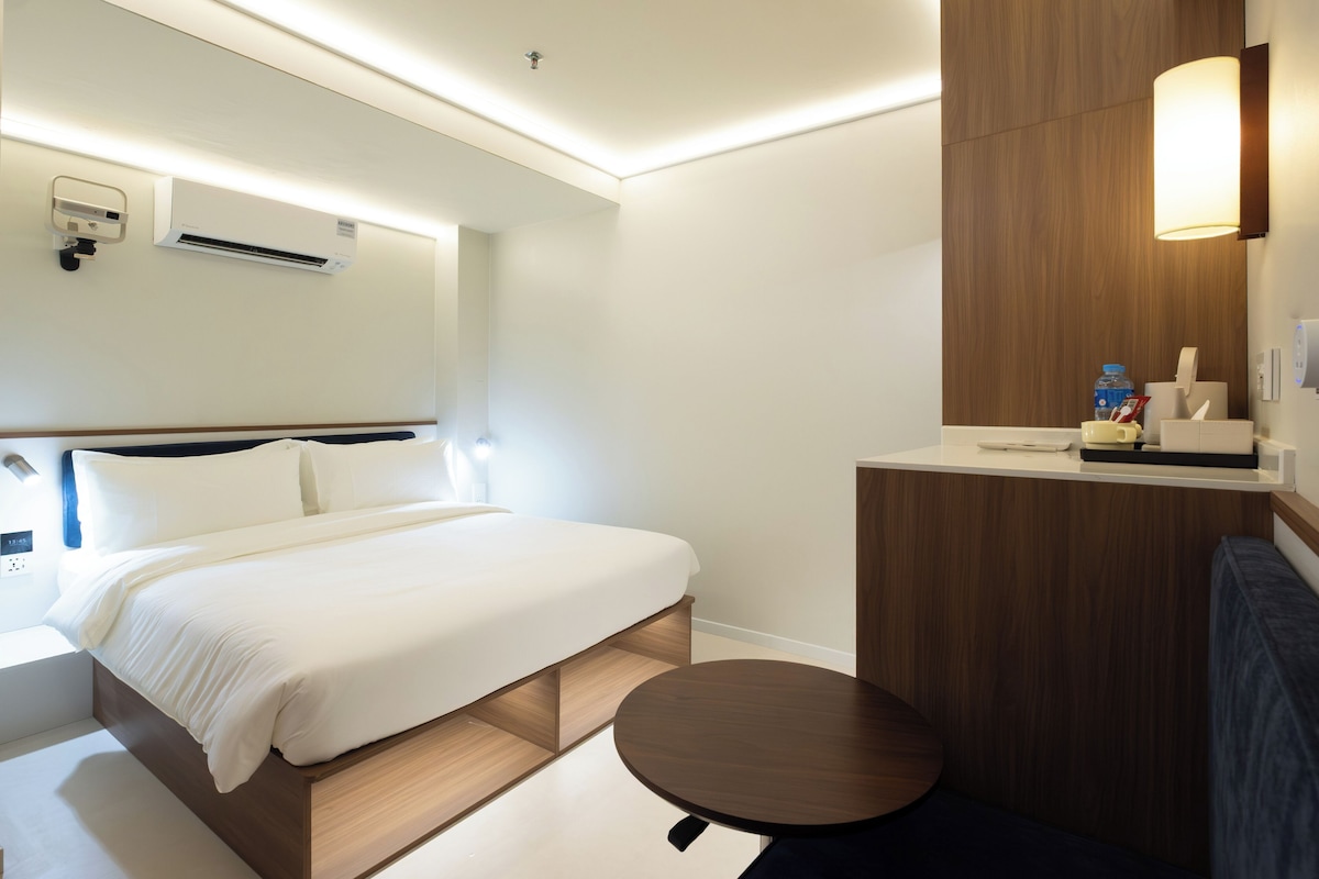 Habitación de hotel con cama, mesita de noche, mesa redonda y espejo con panel de madera.