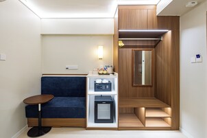 A compact hotel room with a sofa, a minibar, a mirror, and a small table.