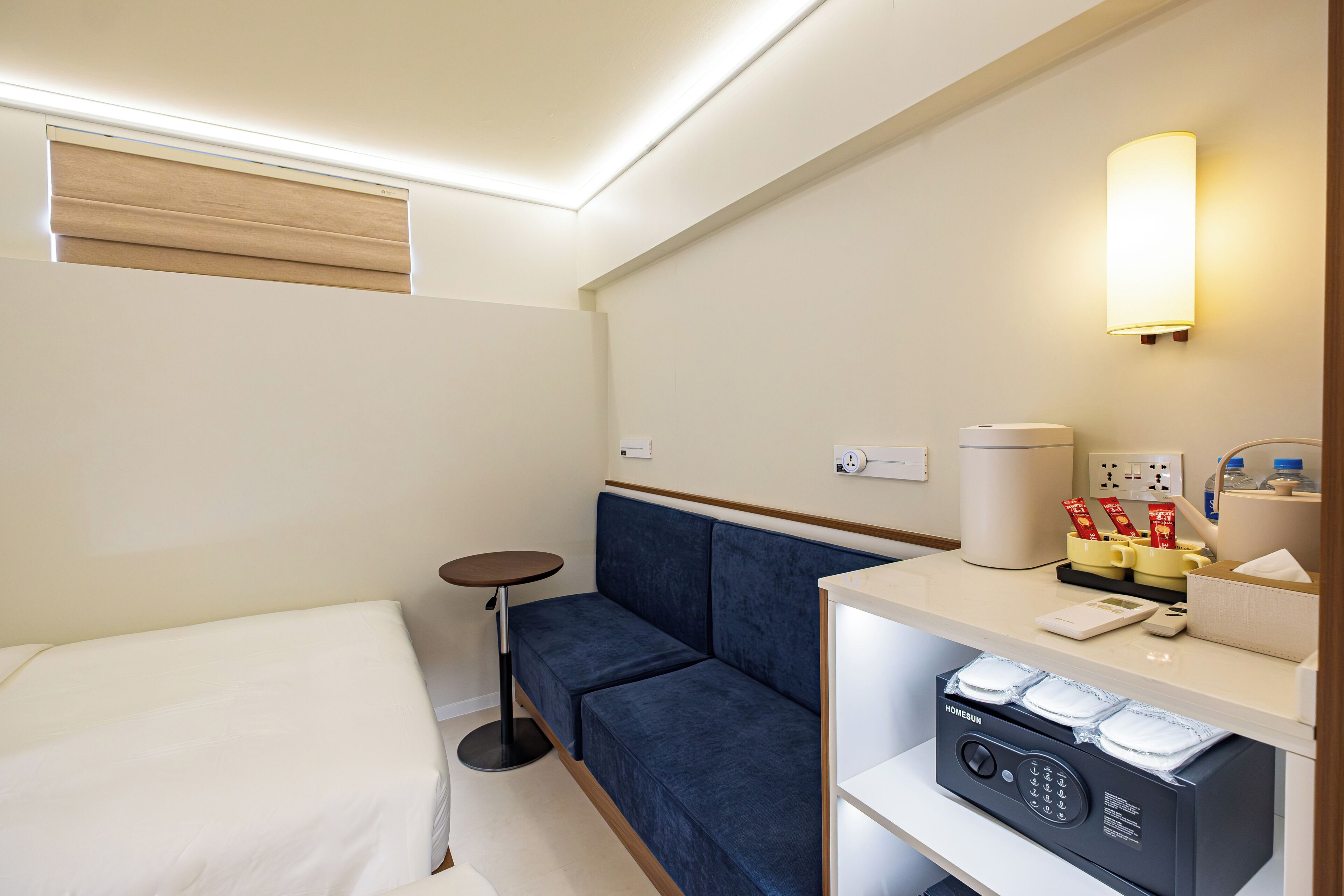 A hotel room with a bed, a blue sofa, a small round table, a minibar, and a wall-mounted lamp.