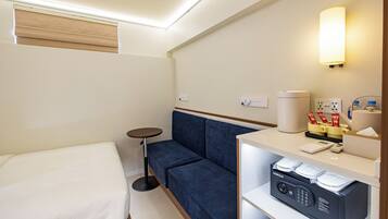 A hotel room with a bed, a blue sofa, a small round table, a minibar, and a wall-mounted lamp.