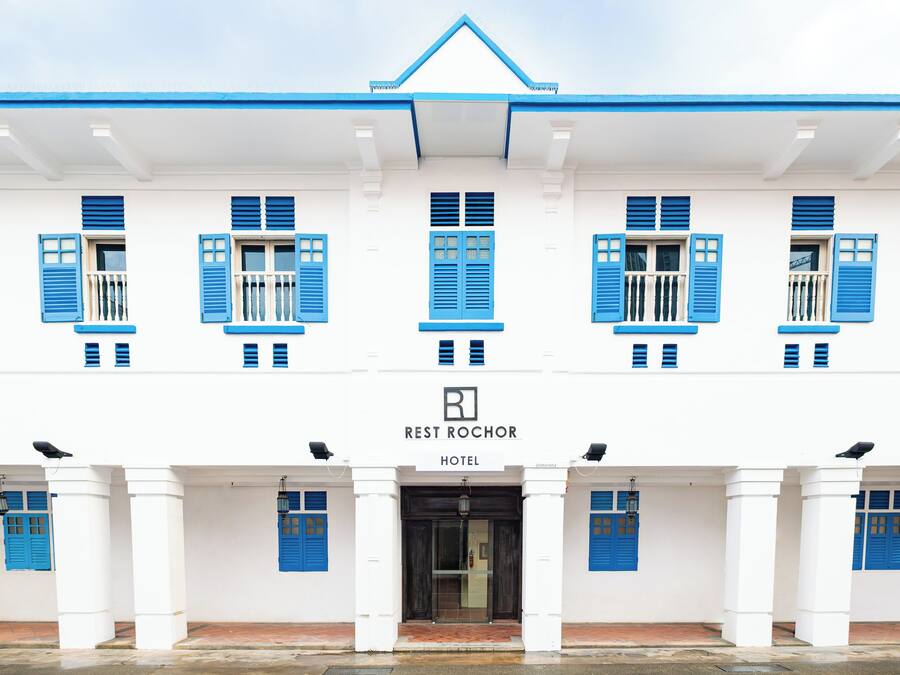 A hotel facade with a symmetrical design, featuring a central entrance, multiple windows with blue shutters, and a sign indicating it is the Rest Rochor Hotel.