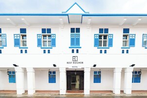A hotel facade with a symmetrical design, featuring a central entrance, multiple windows with blue shutters, and a sign indicating it is the Rest Rochor Hotel.