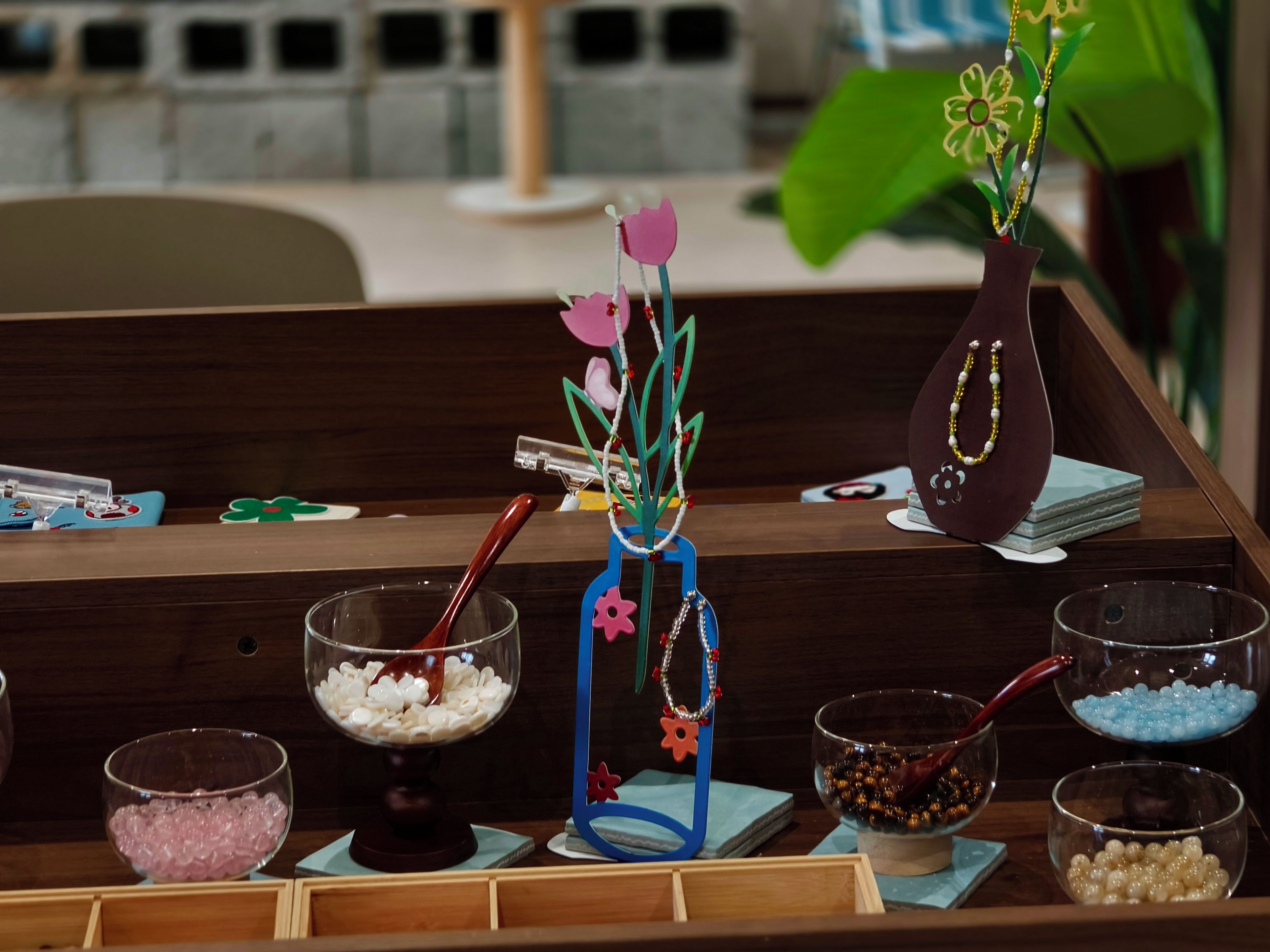 A wooden counter with various bowls containing different colored beads, a decorative vase with a flower, and a small plant.