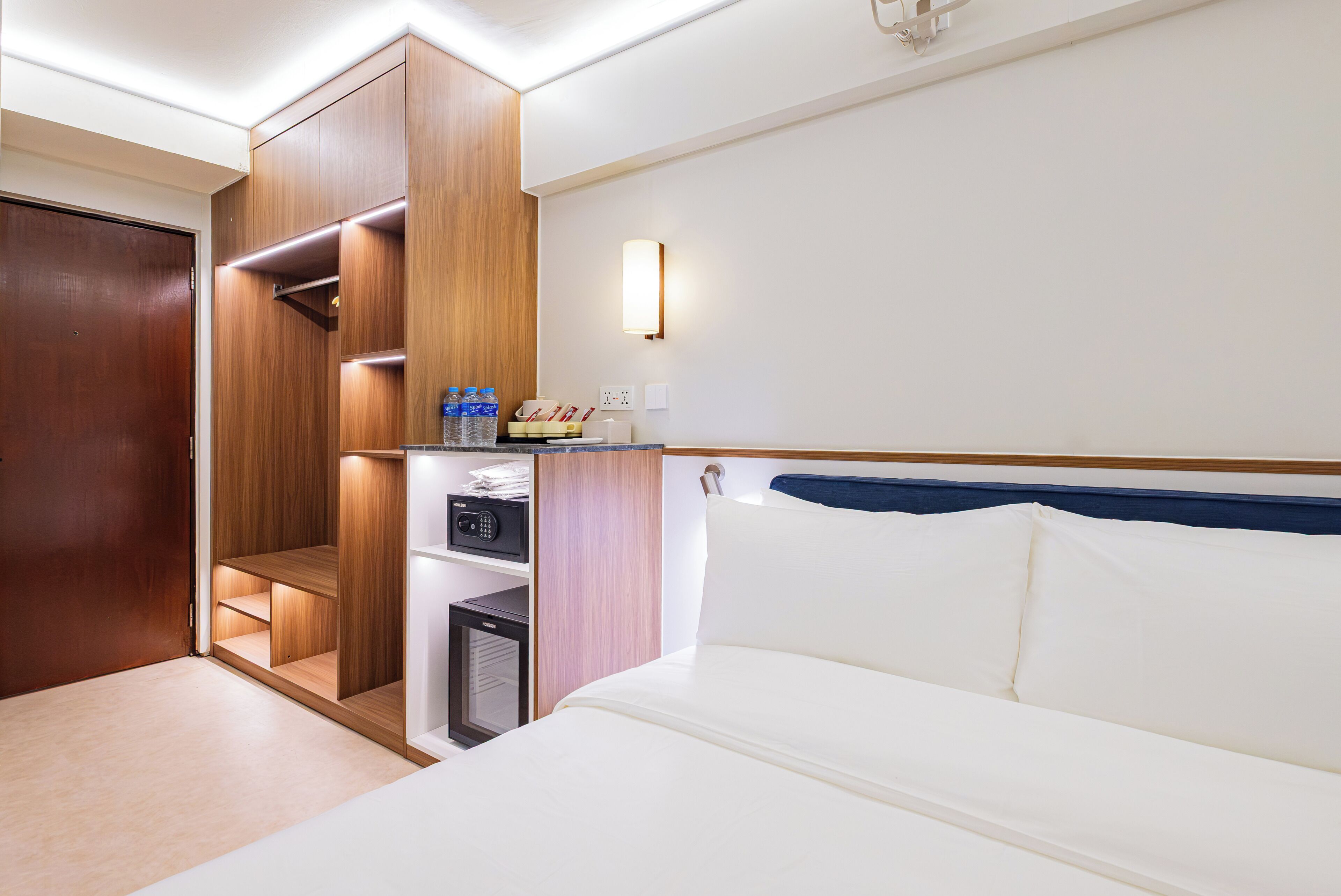 A hotel room with a bed, a wardrobe, a minibar, and a wall-mounted light fixture.