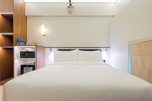 A hotel room with a bed, a nightstand, a shelf with toiletries, and a wall-mounted light fixture.