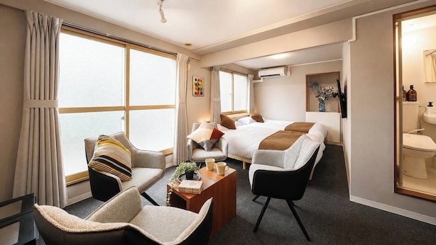 Una habitación de hotel con una cama, dos sillones, una mesa de centro y un amplio ventanal con cortinas.
