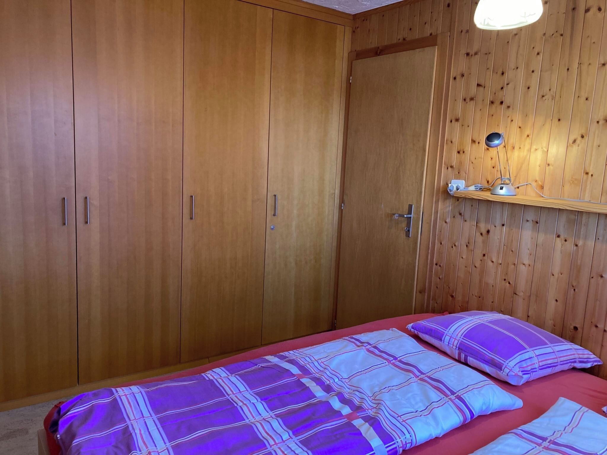 A bedroom with a wooden wardrobe, a bedside table with a lamp, and a bed with patterned bedding.