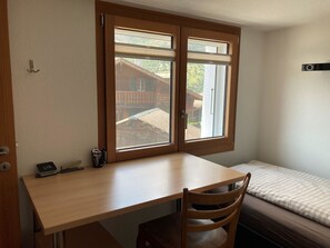 A room with a wooden desk, a chair, a bed with a checkered bedspread, and a window with a view of a wooden building. - Appartement Binna 4-Bettwohnung, Dusche, WC 3-Bettwohnung, Dusche, WC by Interhome (Rosswald)
