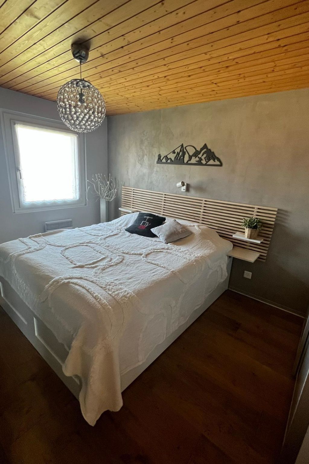 Une chambre avec un plafond en bois, un grand lit recouvert d’une courtepointe blanche, un luminaire fixé au mur et une œuvre d’art représentant une montagne.