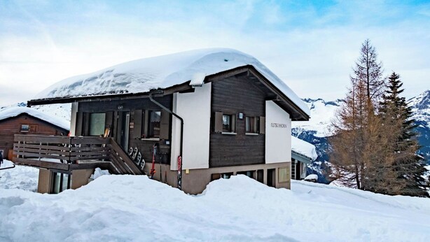 Ein mit Schnee bedecktes Chalet mit einer Holztreppe zum Eingang, einer Seilbahn und einem Berg im Hintergrund.
