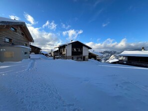 En snedækket landsby med traditionelle træhuse, en klar himmel og et bjerg i baggrunden.