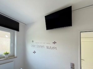 A room with a mounted television, a window with a view of the outside, and a wall with a motivational quote. - "Sandkorn 1" Apartment (Scharbeutz)