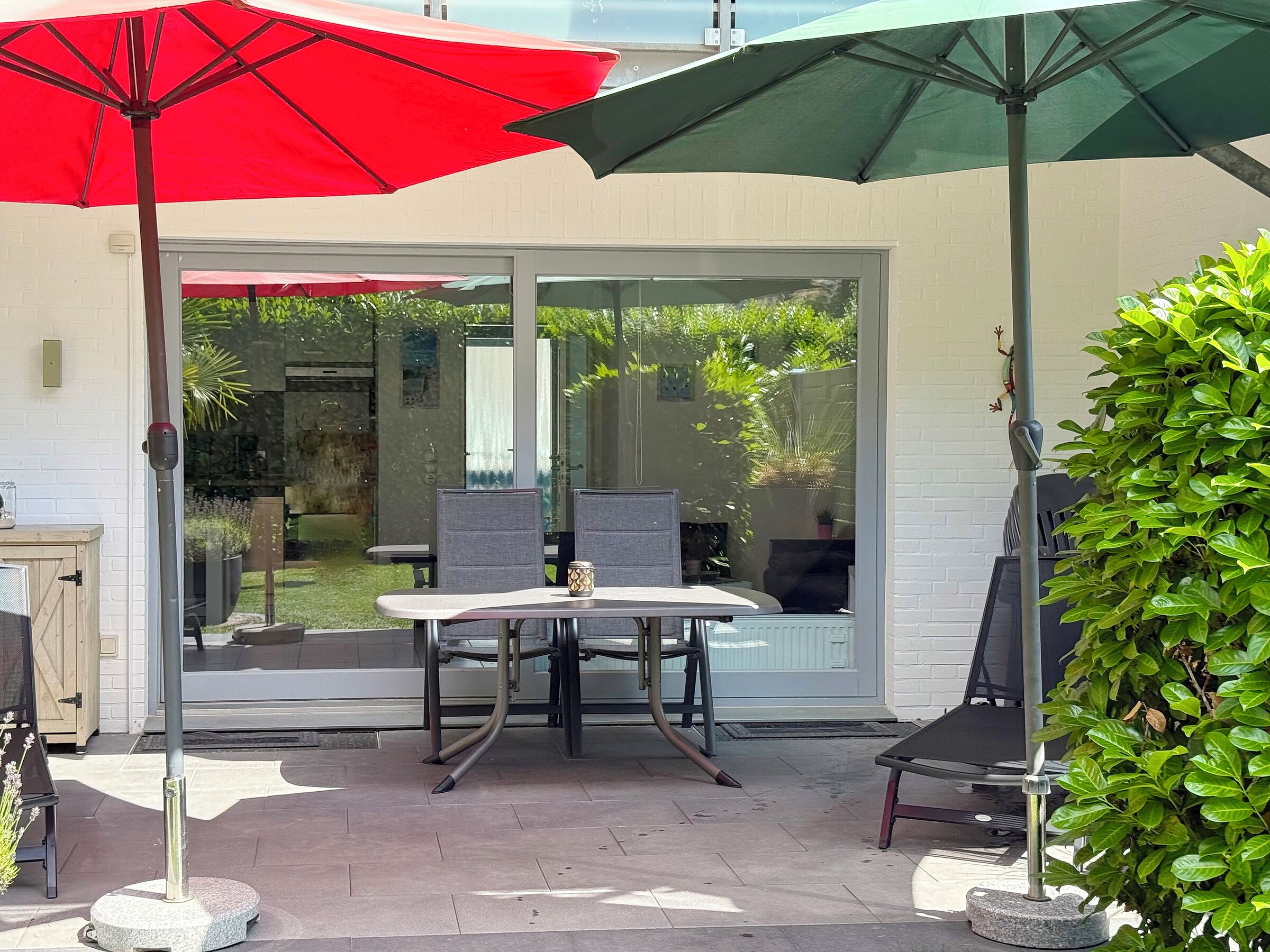 Eine Terrasse mit Tisch und Stühlen, einem roten und grünen Sonnenschirm sowie einem Blumentopf.