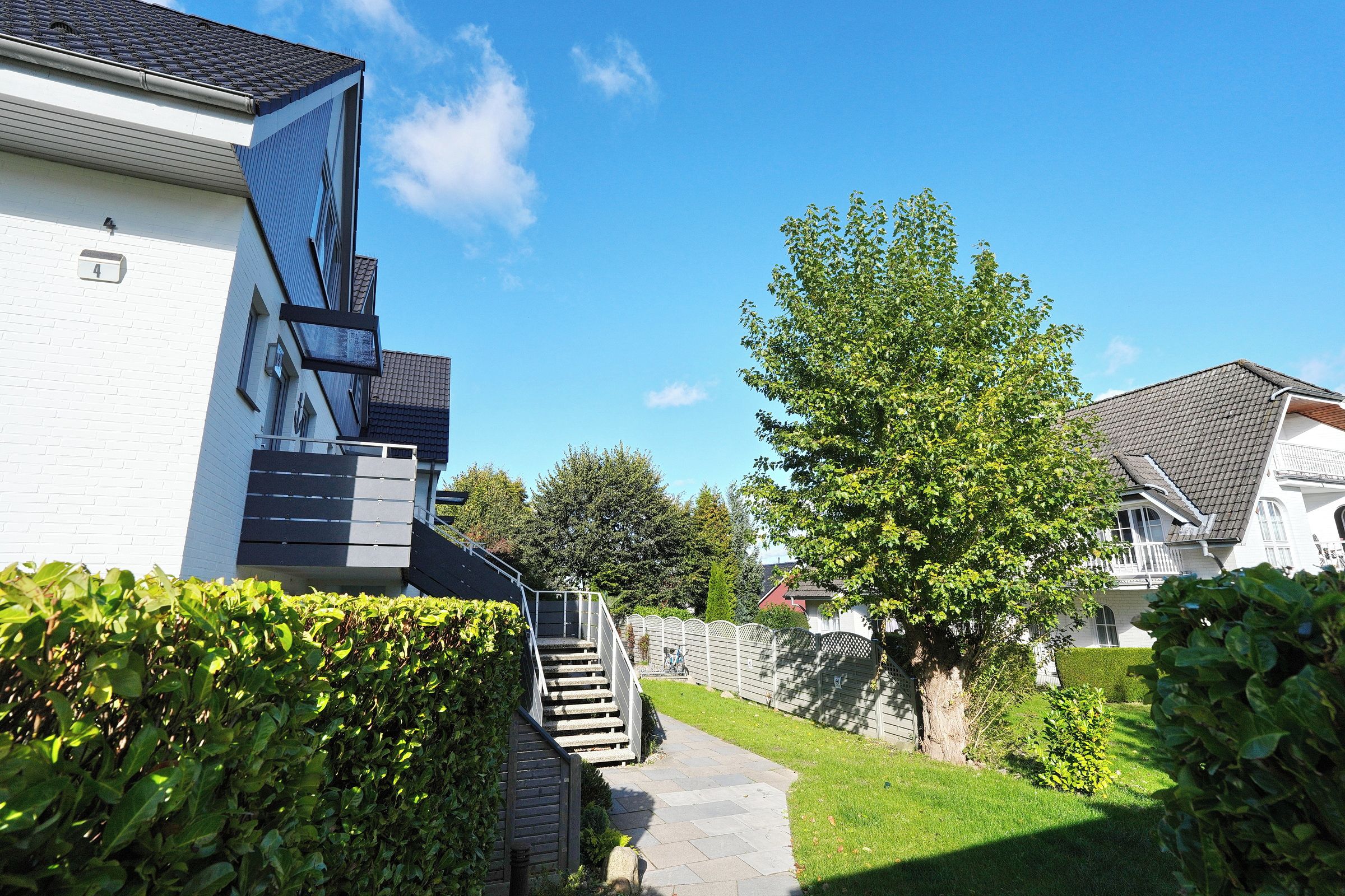 Un quartier résidentiel avec un chemin menant à une maison dont on aperçoit l’escalier et un arbre au premier plan.