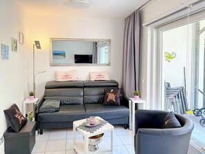 A modern living room with a sofa, a coffee table, and a chair. There is a large mirror above the sofa and a window with curtains. - "Sandkorn 1" Apartment (Scharbeutz)