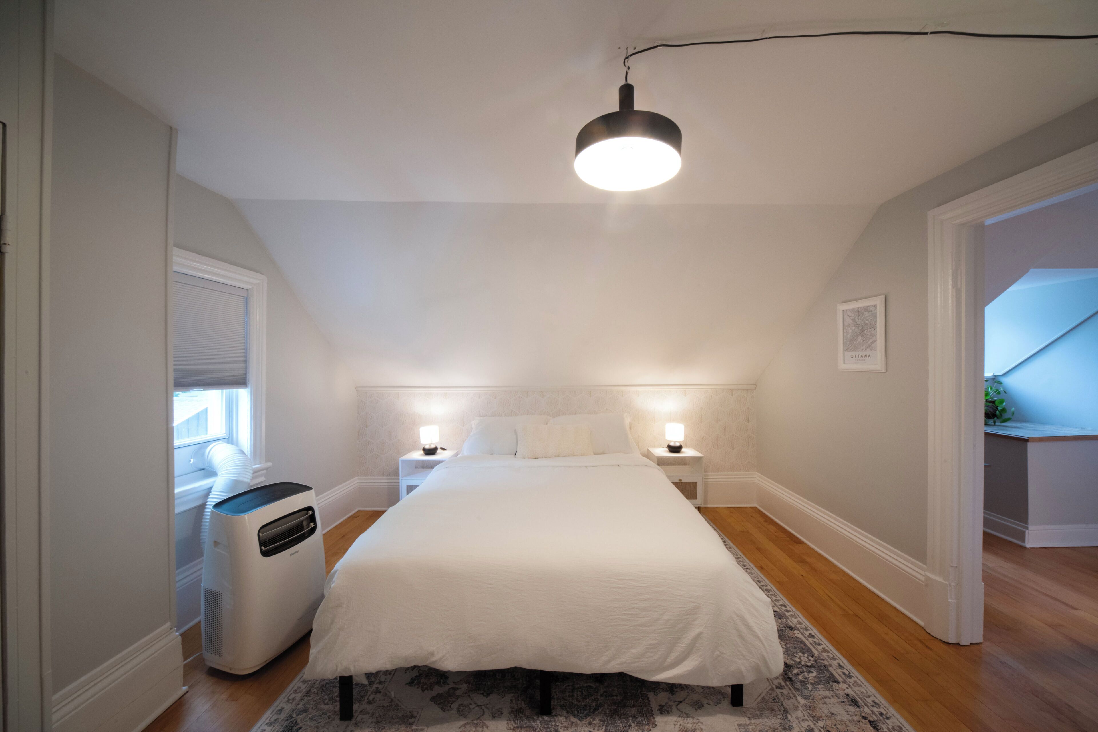 A bedroom with a large bed, two bedside tables with lamps, a ceiling light, a radiator, and a window with blinds.