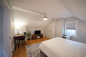 A bedroom with a bed, a desk with a chair, a television, a plant, and a window with blinds.