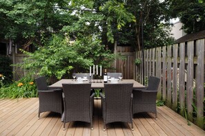 An outdoor dining area with a wooden deck, wicker chairs, a table set with white plates, glasses, and a bottle of wine.