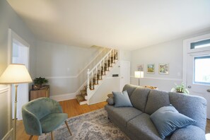 A living room with a grey sofa, a blue armchair, a wooden staircase, and a door with framed pictures.