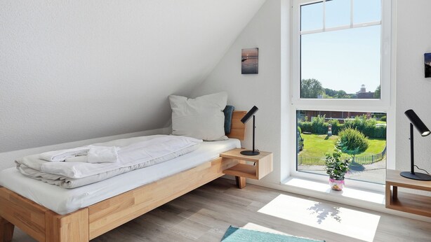 A bedroom with a wooden bed, a small bedside table, a nightstand, a window with a view of a garden, and a wall-mounted picture. - Modern "Ebbe&Flut" Vacation Rental in Norddeich for the Whole Family (Norddeich)