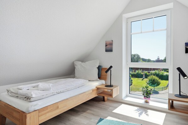 Ein Schlafzimmer mit einem hölzernen Bett, einem kleinen Nachttisch, einem Fenster mit Gartenblick und einem an der Wand hängenden Bild.