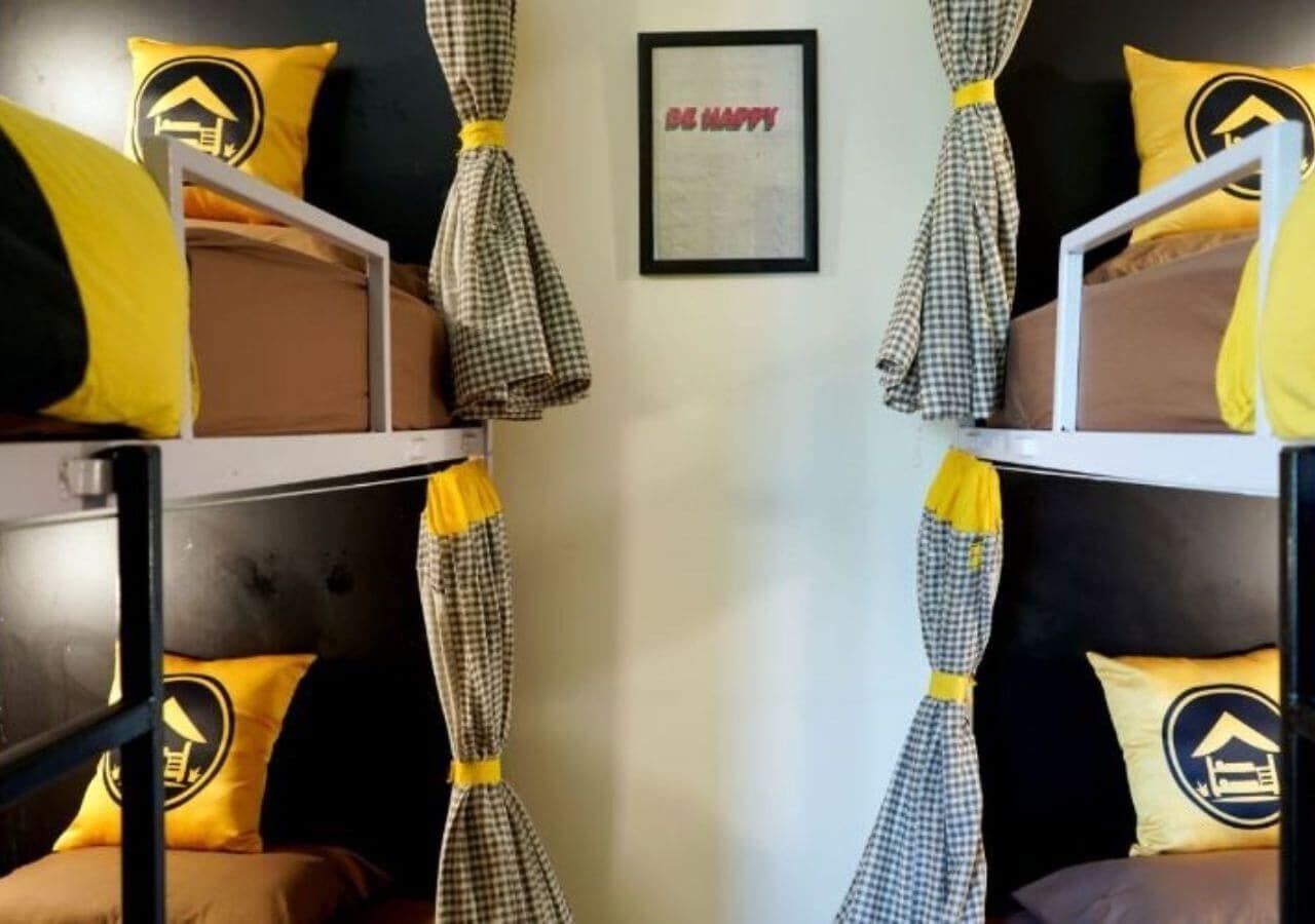 A bunk bed room with checkered curtains tied at the ends, a framed 'BE HAPPY' sign, and yellow pillows with a building logo.