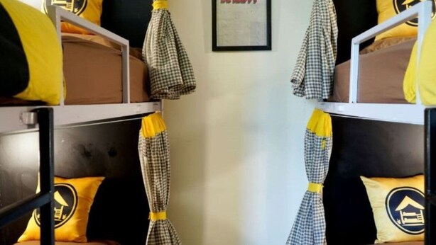 A bunk bed room with checkered curtains tied at the ends, a framed 'BE HAPPY' sign, and yellow pillows with a building logo.