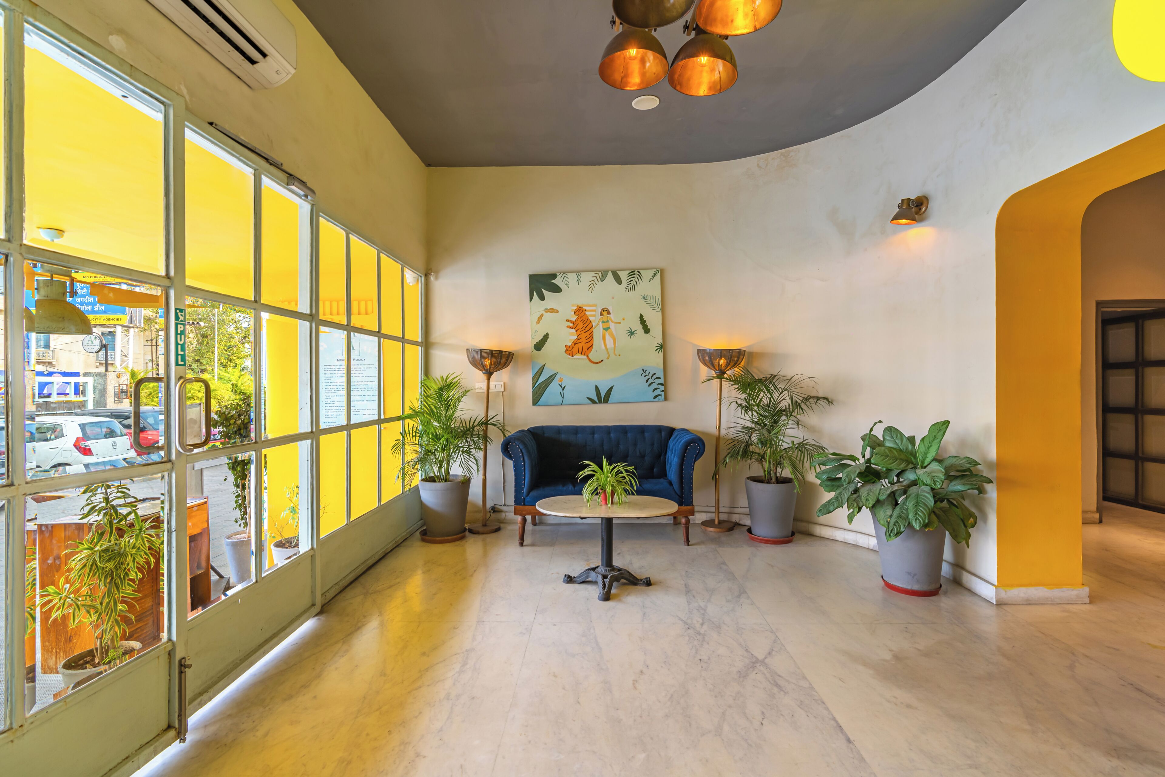 A spacious lobby with a blue sofa, potted plants, and a large window offering a view of the street.