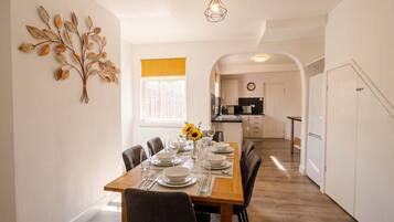 A dining area with a wooden table set for four, a kitchen in the background, and a decorative wall piece.