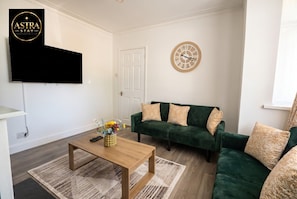 A modern living room with a flat-screen TV mounted on the wall, a green sofa with patterned cushions, a wooden coffee table with a vase of flowers, and a wall clock. - 3-Bed House Sleeps 5 · Free Parking · Walk to Southampton Common (Shirley)