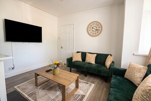 Ein Wohnzimmer mit Flachbildfernseher, einem grünen Sofagarnitur, einem Holztisch und einer dekorativen Wanduhr.