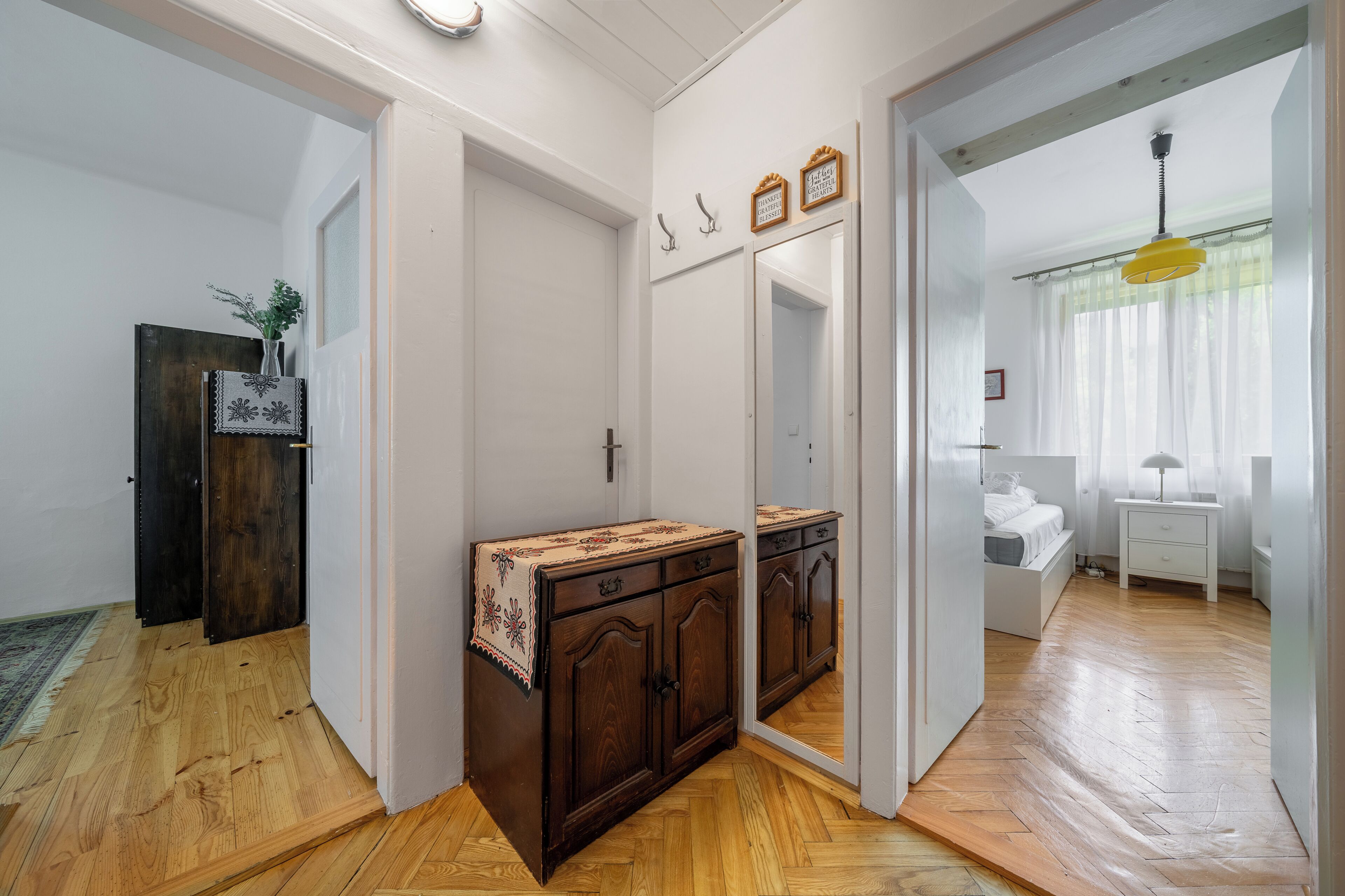 A room with a wooden floor, a large mirror, a wooden cabinet, and a bed with a white headboard.