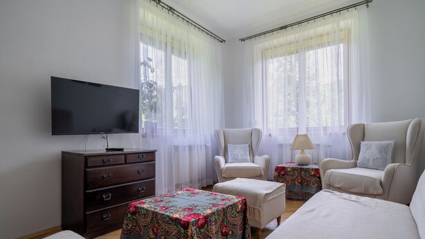 Ein Hotelzimmer mit Flachbildfernseher, einem Holzkoffer, zwei Sesseln, einem kleinen Tisch mit Lampe und einer Tischläufer mit Blumenmuster.