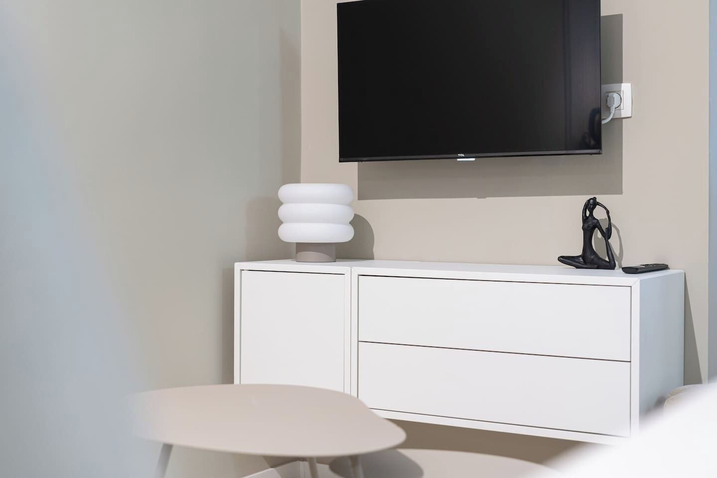 Quarto moderno com um armário branco, uma mesa redonda, uma televisão na parede e uma escultura.
