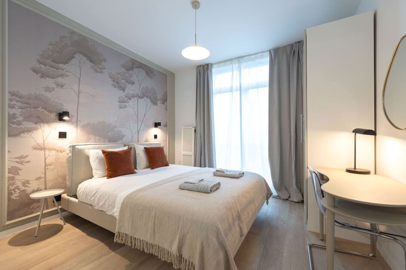 A modern hotel room with a large bed, a desk, and a chair. There is a window with curtains and a wall with a tree mural.