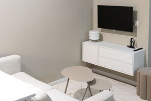Ein modernes Hotelzimmer mit einem flachbildfernseher an der Wand, einer weißen Kommode mit Schubladen, einem kleinen runden Tisch und einem Sofa.