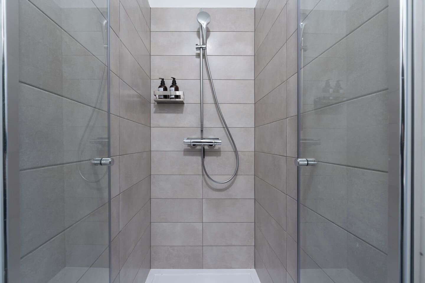 A walk-in shower with a handheld showerhead, a wall-mounted faucet, and a soap dispenser.