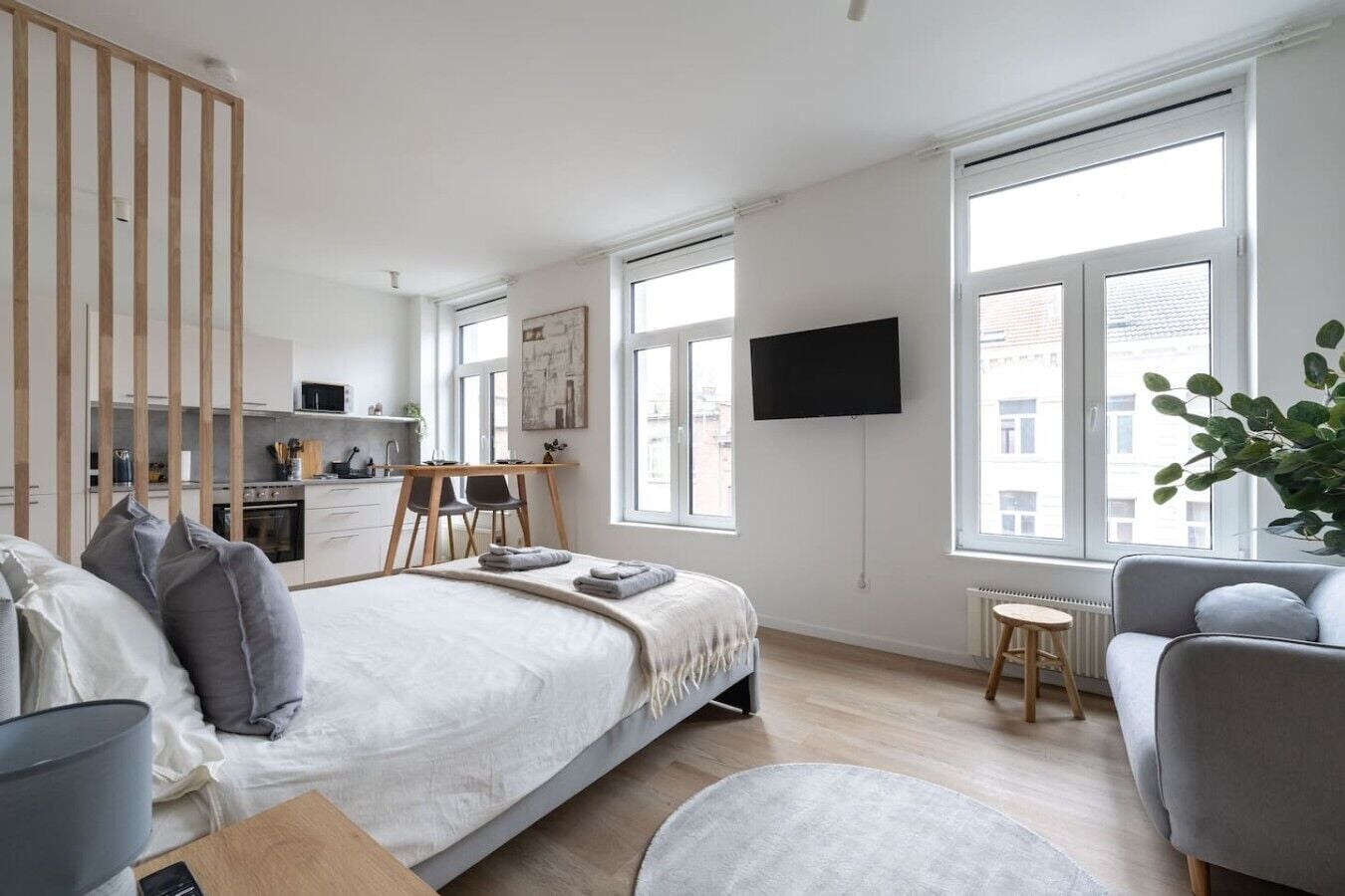 A modern bedroom with a bed, bedside tables, a desk, and a chair. There are large windows providing natural light and a view of the outside.