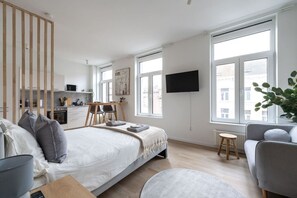 Quarto moderno com cama, mesas de cabeceira, escrivaninha e cadeira. Grandes janelas oferecem luz natural e vista para o exterior.