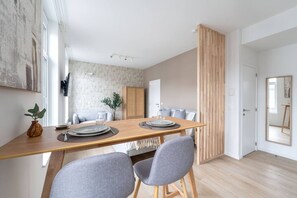 A modern dining area with a wooden table set for two, grey upholstered chairs, a cabinet, and a large window providing natural light. - Modern Cocoon - Gare du Midi - BRO144.2.1 (Anderlecht)