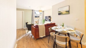 Un salon moderne avec un canapé, une table à manger et des chaises. Il y a une fenêtre avec des rideaux, une plante et un tableau au mur.