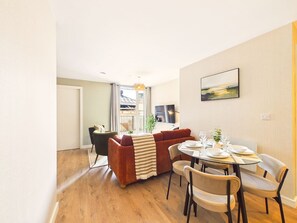 Una sala de estar moderna con un sofá, una mesa de comedor y sillas. Hay una ventana con cortinas, una planta y un cuadro en la pared.