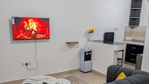 A living room with a sofa, a coffee table, a TV displaying a fireplace, a small kitchen area with a fridge, and a water dispenser. - Perfect apartment in Nairobi for a relaxing stay (Nairobi)