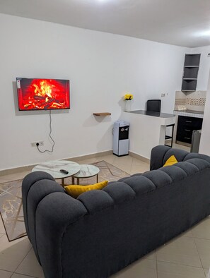 Ein Wohnzimmer mit einer Couch, einem Couchtisch, einem Fernseher, der einen Kamin zeigt, einer kleinen Küchenzeile mit Kühlschrank und Wasserspender.