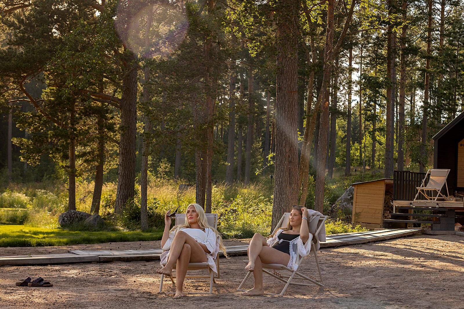 Dos personas sentadas en sillas al aire libre en un área boscosa, con una cabaña de madera al fondo.
